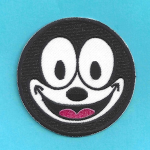 May include: A circular embroidered patch featuring a cartoon cat face with large white eyes, a black nose, and a wide, smiling mouth with a pink tongue. The patch has a black border and is set against a light blue background.