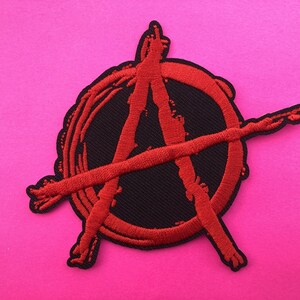 May include: Embroidered patch displaying the anarchy symbol, a red "A" inside a circle, with a line through it. The symbol is stitched in red thread on a black base. The patch has a scalloped edge and is set against a bright pink background.