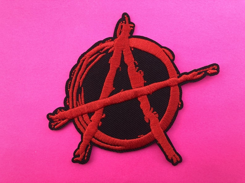 May include: Embroidered patch displaying the anarchy symbol, a red "A" inside a circle, with a line through it. The symbol is stitched in red thread on a black base. The patch has a scalloped edge and is set against a bright pink background.