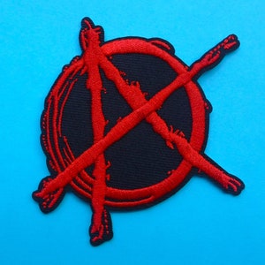 Puede incluir: Un parche negro con un símbolo de anarquía rojo. El símbolo es un círculo con una A mayúscula y líneas cruzadas, bordado con hilo rojo. El parche está sobre un fondo azul brillante. El parche tiene forma circular.