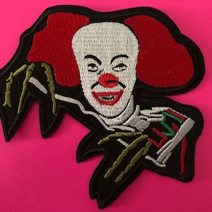 May include: Embroidered patch depicting a scary clown with a white face, red hair, and a red nose and mouth. The clown has black arms with green claws and is wearing a black outfit. The patch is on a pink background.