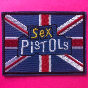 Könnte beinhalten: Gestickter Aufnäher mit dem Union Jack-Design und den Worten "Sex Pistols". Der Aufnäher hat einen blauen Hintergrund mit roten und weißen Streifen und einen schwarzen Rand. Der Text "Sex" ist gelb und "Pistols" weiß. Der Aufnäher befindet sich auf einem rosa Hintergrund.