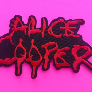 May include: Embroidered patch with the name "ALICE COOPER" in red, stylized lettering on a black background. The patch has a unique, irregular shape and is set against a bright pink backdrop.