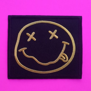 Può includere: Toppa rettangolare nera con un ricamo dorato a forma di faccina sorridente. La faccina ha gli occhi a croce e la lingua di fuori. La toppa è su uno sfondo rosa acceso.