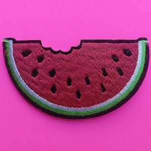 May include: Embroidered watermelon slice patch with a bite taken out. The patch features a red interior with black seeds, a green and light blue rind, and a black border. The patch is on a pink background.