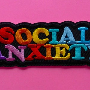 Peut inclure: Écusson brodé avec le texte "SOCIAL ANXIETY" en fil multicolore. Les lettres sont bordées de noir et se détachent sur un fond rose. Le patch a une bordure noire.