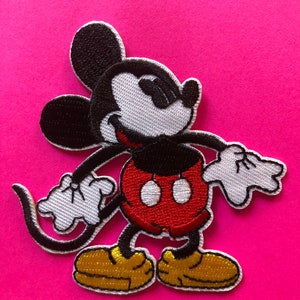 Könnte beinhalten: Gestickter Aufnäher mit Mickey Mouse in einer klassischen Pose. Der Aufnäher zeigt Mickeys ikonische schwarzen Ohren, schwarzen Körper, rote Shorts, gelbe Schuhe und weiße Handschuhe. Der Aufnäher befindet sich auf einem leuchtend pinkfarbenen Hintergrund.