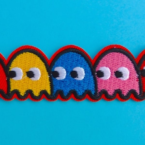 Può includere: Cinque toppe ricamate a forma di fantasmi rossi, gialli, blu e rosa. Ogni fantasma ha occhi bianchi con pupille nere e un contorno nero. Su sfondo blu brillante.