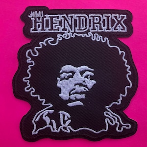 Puede incluir: Parche bordado negro con un retrato de Jimi Hendrix con costuras blancas. El parche tiene el nombre "Jimi Hendrix" bordado en blanco en la parte superior. El parche está sobre un fondo rosa.