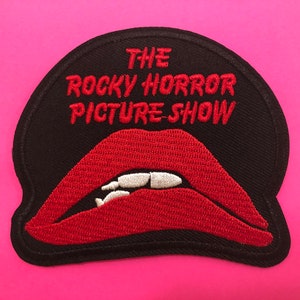 May include: Embroidered patch featuring the text "THE ROCKY HORROR PICTURE SHOW" in red on a black background. Below the text is a red, open-mouthed design with white teeth. The patch is on a pink background.
