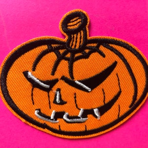 May include: Embroidered orange pumpkin patch with a black outline and stem. The pumpkin has a carved face with black eyes and mouth, and white stitching details. The patch is on a pink background.