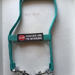 Może przedstawiać: Turkusowa smycz dla psa z czarnym, wyściełanym uchwytem i dwoma metalowymi klipsami. Czarno-biała naszywka z napisem "STOP IGNORE ME I'M WORKING". Smycz jest przeznaczona dla właścicieli zwierząt.