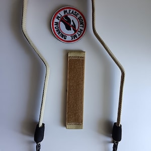 Op de afbeelding: Een beige hondenriem met een zwart gevoerd handvat en twee zwarte metalen clips. Inbegrepen is een ronde witte patch met een rode rand en zwarte tekst die luidt PLEASE DONT PET ME I'M WORKING, en een rechthoekige beige klittenbandstrip.