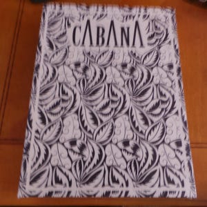 May include: A rectangular book or box cover featuring a black and white repeating leaf pattern. The word CABANA is printed in a bold, stylized black font at the top center. The design is enclosed within a thin black border on a white background.