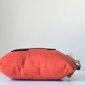 May include: A red pillow with black squares on the front. The pillow is made of a textured fabric and has a brown fringe on the side.