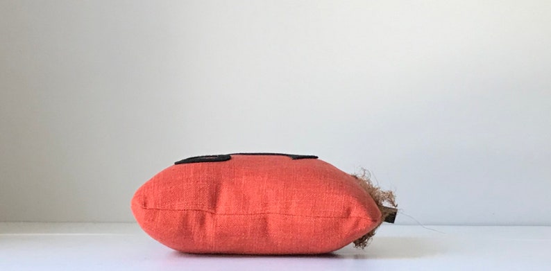 May include: A red pillow with black squares on the front. The pillow is made of a textured fabric and has a brown fringe on the side.