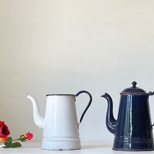 May include: Two vintage enamelware coffee pots, one white and one navy blue, with black trim and handles. A red rose with green leaves is in the foreground. The pots have a classic, old-fashioned design.