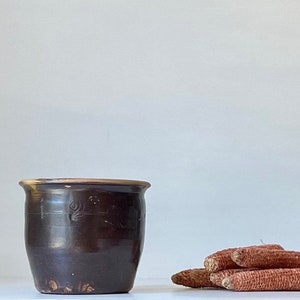 May include: A dark brown ceramic pot with a decorative design sits on a white surface next to a pile of dried red corn cobs.