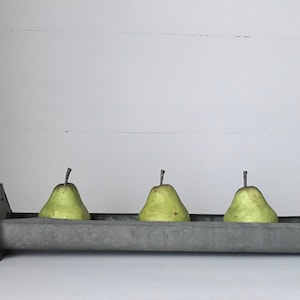 May include: A galvanized metal trough with four green pears inside. The trough is sitting on a white surface.
