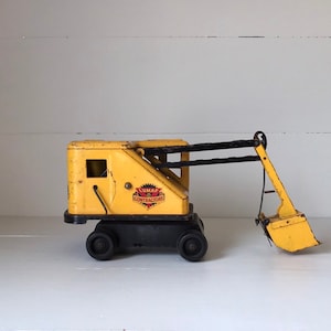 May include: Yellow toy construction crane with a black arm and a yellow bucket. The crane has a black base and wheels. The side of the crane says "Lumar Contractors".