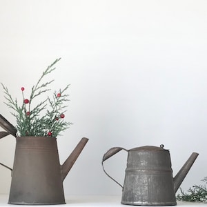 May include: Two vintage metal teapots, one with a sprig of greenery and red berries, the other with a sprig of greenery. The teapots are both in a rustic, aged style.
