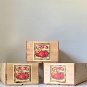 May include: Three wooden boxes stacked on top of each other. Each box has a label with the text "Faultless" and a picture of red apples.