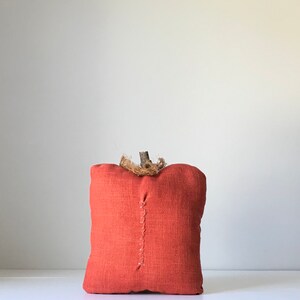 May include: A small, orange fabric pumpkin with a twig stem and burlap accents. The pumpkin is sitting on a white surface.