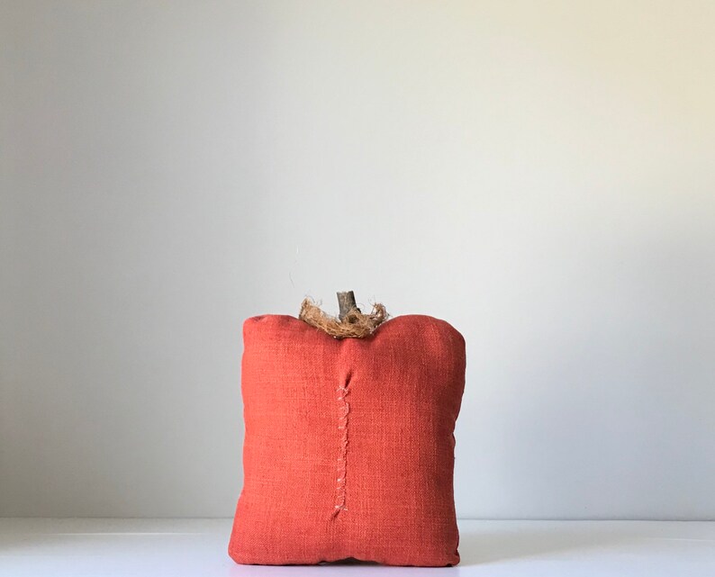 May include: A small, orange fabric pumpkin with a twig stem and burlap accents. The pumpkin is sitting on a white surface.