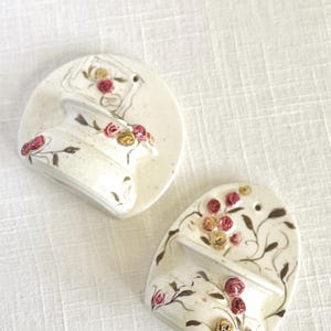 May include: Two off-white ceramic wall pockets with floral designs. Each pocket features a small shelf and is decorated with red and yellow roses and brown vines. The pockets are on a white textured surface.