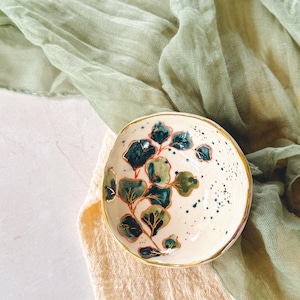 May include: A small, off-white ceramic bowl with a gold rim. The bowl is decorated with a botanical design featuring green and blue leaves. The bowl is resting on a beige cloth, with a green cloth in the background.