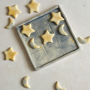 May include: A ceramic tic-tac-toe game with a celestial theme. The game board is a square with nine blue squares. The game pieces are star and crescent moon shapes in shades of yellow and white.