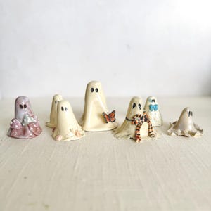 May include: A collection of seven small ceramic ghost figurines with painted black eyes. One ghost features a butterfly, another a striped bow, one has mushrooms, and another a blue bow. Each ghost has a smooth, glossy finish in various shades.
