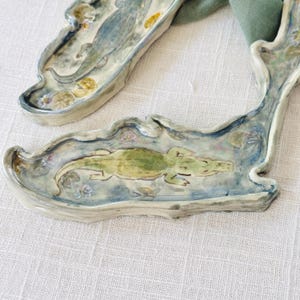 May include: Two ceramic alligator-shaped serving dishes. The dishes are painted with a green alligator in a blue and white water scene, with lily pads. The edges are scalloped and have a light gray color.