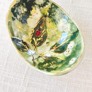 May include: Oval-shaped ceramic dish with a nature-inspired design. The dish features a green and yellow background with gold accents. A red ladybug sits on a leaf with gold veins. The rim is trimmed with gold.