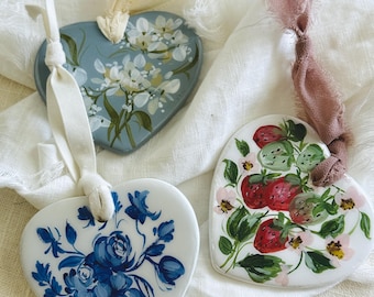 Op de afbeelding: Drie hartvormige ornamenten met handgeschilderde bloemmotieven. Eén is lichtblauw met witte bloemen, een andere met blauwe rozen en de derde met aardbeien. Elk heeft een lint om op te hangen.