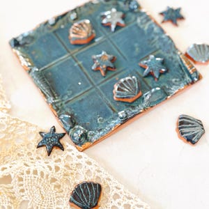 May include: A handmade ceramic tic-tac-toe game set. The game board is a square with a blue glaze and a grid pattern. The game pieces are starfish and seashell shapes in blue and brown. The set is on a white surface with a lace cloth.