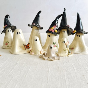 May include: A collection of nine ceramic ghost figurines in various sizes, some wearing black pointed hats. Several ghosts feature hand-painted floral details in pink, yellow, and green, while one ghost has a small orange butterfly on its side.