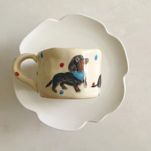 Puede incluir: Una taza de cerámica color crema con un perro salchicha negro y marrón pintado a mano que lleva un pañuelo azul, acentuado con pequeños puntos rojos y azules, sobre un platillo blanco festoneado con un borde curvo y acabado brillante.