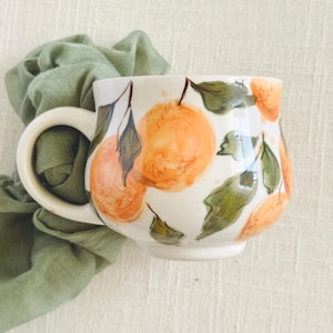 May include: A white ceramic mug with a large handle, decorated with hand-painted orange fruit and green leaves. The mug is set against a light green cloth background, creating a fresh and natural aesthetic. The mug is perfect for enjoying your favorite beverage.