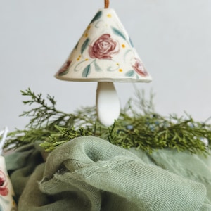May include: A ceramic mushroom-shaped ornament with a floral design in red, green, and yellow. The ornament hangs from a brown string and rests above a green cloth and greenery.
