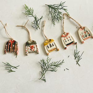 May include: Five ceramic ornaments shaped like miniature sweaters, each featuring unique hand-painted designs including stripes, floral patterns, and pine trees. Each ornament hangs from a thin brown twine loop for display purposes.