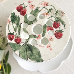 Op de afbeelding: Een ronde keramische bloemenkikker met een gespikkelde crèmekleurige afwerking, voorzien van een handgeschilderd patroon van rode aardbeien, groene bladeren en kleine roze bloemen. Het bovenoppervlak heeft meerdere ronde gaten.
