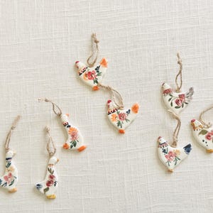 May include: A collection of seven white ceramic ornaments shaped like chickens and geese, each featuring hand-painted floral patterns in red, orange, and blue. Every ornament is suspended by a thin, natural-colored jute twine loop for hanging.