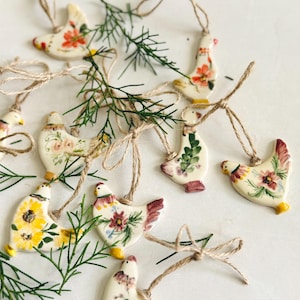 May include: A collection of cream-colored ceramic chicken ornaments, each hand-painted with unique floral patterns in shades of red, yellow, pink, and green. Every ornament features a rustic jute twine loop for hanging and is accented with green pine sprigs.