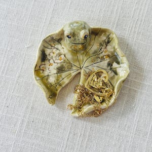 Puede incluir: Una bandeja de cerámica con forma de hoja y cara de rana. La bandeja es principalmente verde con detalles dorados y contiene un collar dorado en forma de corazón y otras joyas doradas. La bandeja está sobre una superficie blanca.