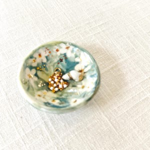 May include: A small, round, ceramic dish with a teal and white floral design. The dish contains a gold-colored charm with white and gold accents and a small, white seashell. The dish is on a white, textured surface.