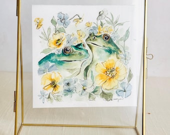 Puede incluir: Una pintura de acuarela de dos ranas verdes rodeadas de flores amarillas y azul claro, encerrada en un marco de metal color latón rectangular con paneles de vidrio transparente. La obra de arte presenta una firma en la esquina inferior derecha.