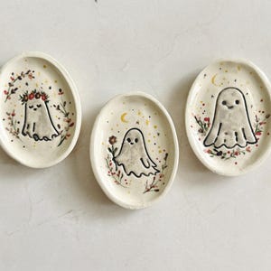 May include: Three oval ceramic trinket dishes in off-white, each featuring a hand-painted ghost character. The ghosts are decorated with small floral wreaths, tiny stars, crescent moons, and delicate botanical accents in red, green, and gold.