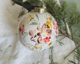 May include: A white ceramic Christmas ornament with a floral design in shades of pink, yellow, and brown. The ornament has a gold-colored metal cap and a green ribbon for hanging. The ornament is resting on a white cloth with green sprigs.