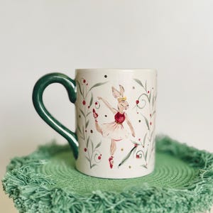 Może przedstawiać: Ceramiczny kubek z zielonym uchwytem i fantazyjnym wzorem. Kubek przedstawia baletnicę-królika w różowej tutu, otoczoną kwiatowymi akcentami. Kubek stoi na zielonej podkładce z frędzlami.
